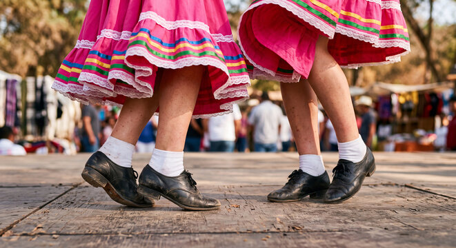 Dancers in colorful traditional folk costumes perform lively steps wearing dancing shoes on an outdoor stone floor.