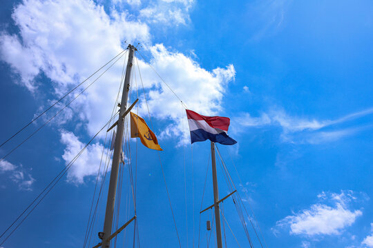 Low angle view of ship masts with a waving Dutch national flag against a bright blue sky with white clouds