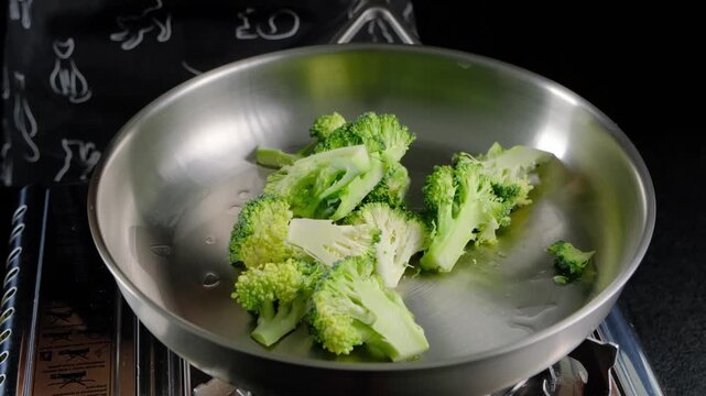 Close-up of hands cooking fresh green broccoli florets in a metal skillet on a stove in the Cooking location, daytime indoor kitchen scene.