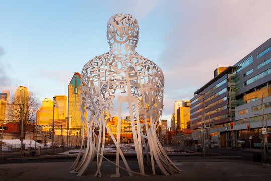 Montreal, Quebec, Canada, March 25, 2025 - Horizontal view of the back of The Source statue by Jaume Plensa at sunrise with downtown skyline in the background, Griffintown sector