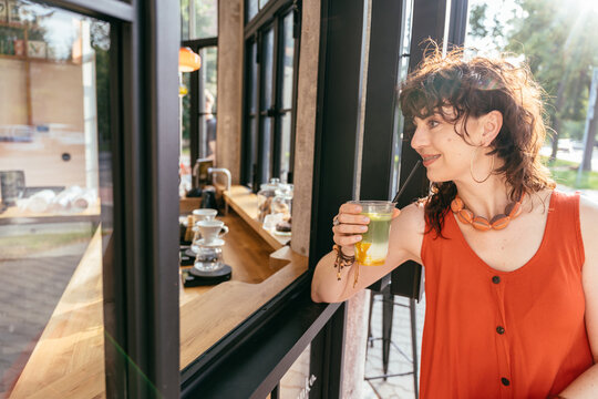 Cheerful middle aged woman in orange dress drinking iced matcha by open modern cafe window