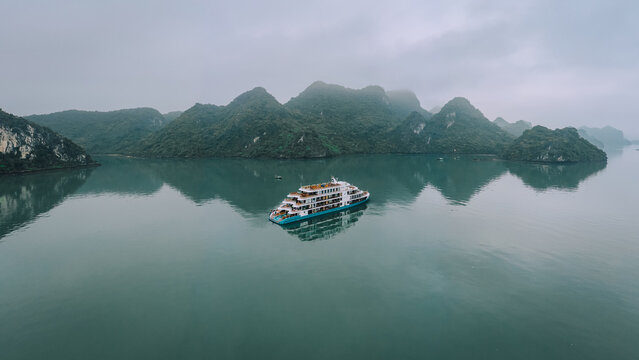 Aerial view of a cruise ship sailing through the calm waters of Ha Long Bay, surrounded by limestone karst mountains under an overcast sky in Ha Tu, Quang Ninh, Vietnam.