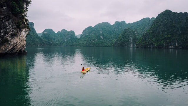 Aerial view of a person kayaking in the calm waters of Ha Long Bay, surrounded by towering limestone karsts and lush green cliffs in Ha Tu, Quang Ninh, Vietnam.