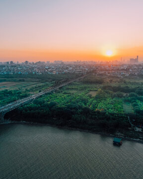 Aerial view of Long Bien Bridge spanning the Red River at sunset, with lush greenery, urban cityscape, and golden light in Hong Ha, Hanoi, Vietnam.