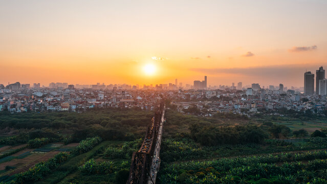 Aerial view of Long Bien Bridge spanning over lush green fields with the urban skyline of Hanoi in the background during a golden sunset, Hanoi, Hanoi, Vietnam.