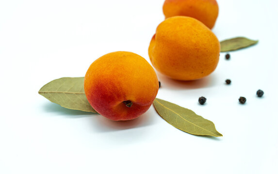 Three apricots with bay leaves and black peppercorns arranged on a white background