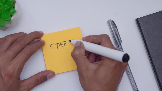 a man writing start now on orange sticky note at his white desk