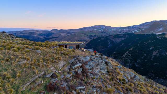 Aerial view of Fallow Ridge Retreat on a rocky cliffside with tussock grass and rolling hills at sunset, Waikouaiti, Otago Region, New Zealand.