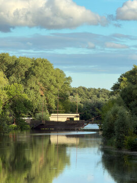 Wooden ship replica on calm river with reflection, historic vessel among green trees