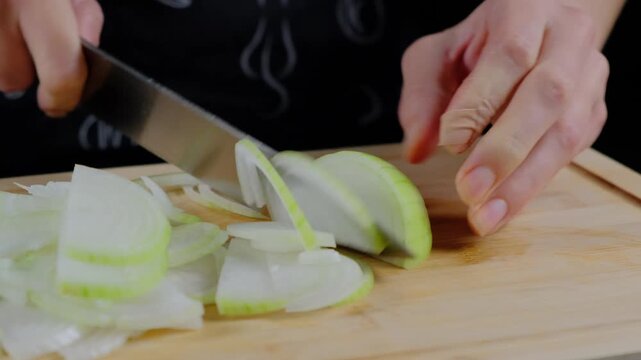 Adult hands use a chef knife to slice onions on a wooden cutting board in a Cooking kitchen, daytime.