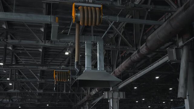 Suspended industrial exhaust hood beside orange hose reels beneath steel truss ceiling inside factory interior with ductwork, pipes, gridded structure, and worn service surfaces