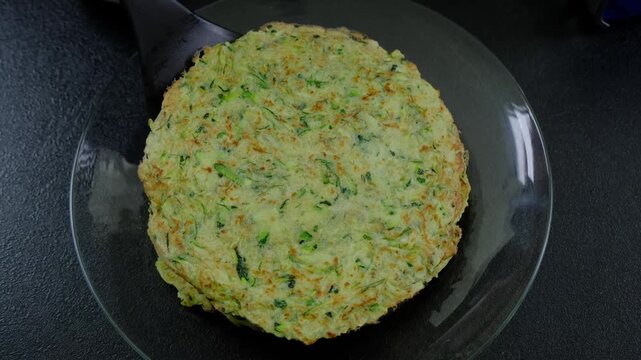 Overhead view of a large green-speckled fried vegetable fritter made of shredded zucchini and spinach on a clear glass plate, set on a dark countertop in a kitchen, during daytime at Cooking location.