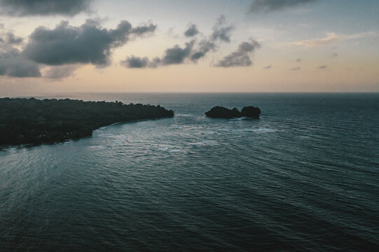 Aerial view of the coastline with a small island in the Caribbean Sea, lush tropical forest, and calm waves under a soft sunset sky in Manzanillo, Limon Province, Costa Rica.