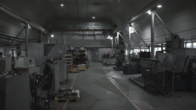 Industrial machine shop interior with central aisle, heavy machinery, metal workbenches, stacked parts, arched ceiling, bright side windows and workers near equipment