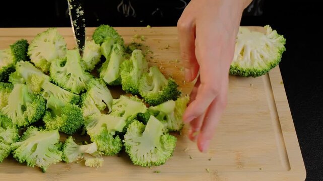 Adult hand uses knife to chop fresh broccoli florets on wooden cutting board in a cooking kitchen during daytime.
