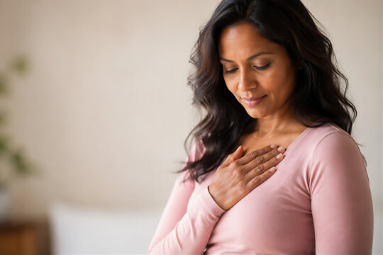 Woman with hand on heart in blush pink top looking down with peaceful expression, self-care and women health awareness concept