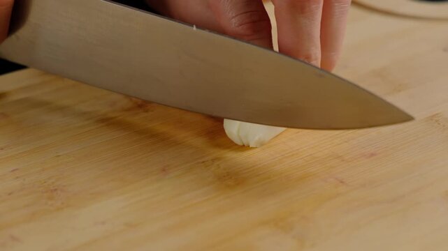 Close-up of hands chopping garlic cloves with a knife on a wooden board during food preparation in a Cooking kitchen, daytime.