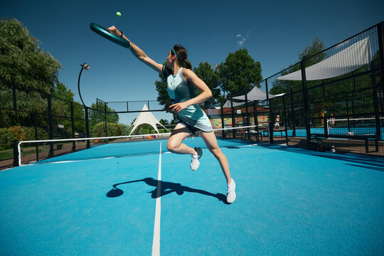 Stylish female padel player in blue activewear extending for high ball on sunny outdoor court. Concept of womens athletic fashion, functional sportswear, energetic brand campaign.