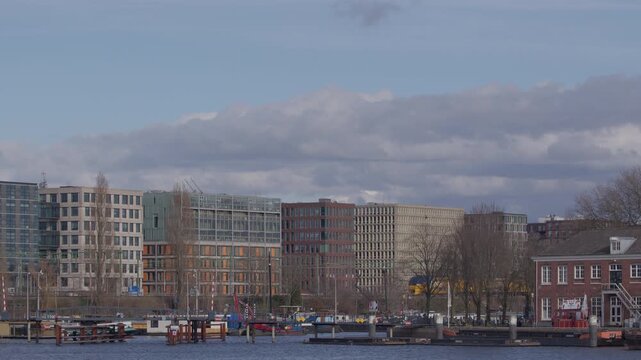 A wide landscape view of Amsterdam's modern waterfront architecture along the IJ bay, featuring a yellow Dutch NS passenger train passing by office buildings and residential blocks on an overcast day.