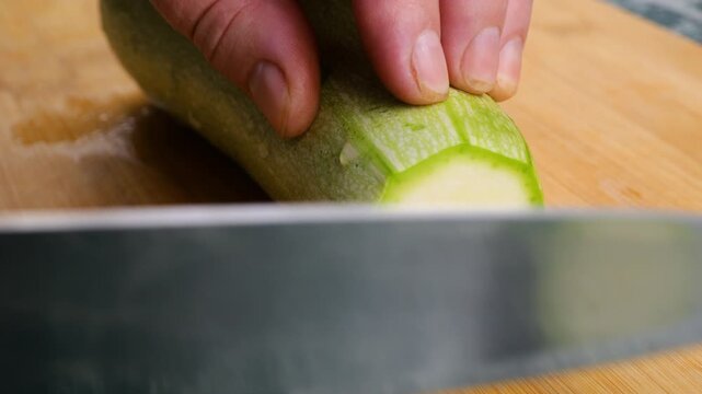 A chef uses a sharp knife to slice a fresh green zucchini into smooth round pieces on a wooden cutting board, preparing healthy organic ingredients for a home meal.