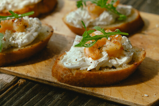 Bruschetta or crostini with cream cheese, shrimp and arugula on wooden cutting board. Tasty healthy snacks.