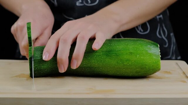 A woman in a black apron slices a large green zucchini on a wooden cutting board in a kitchen during daytime, Cooking location.