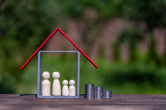Family budget and real estate mortgage investment concept. Wooden peg doll family inside house frame and stacked coins against green yard backdrop, representing property savings finance and insurance