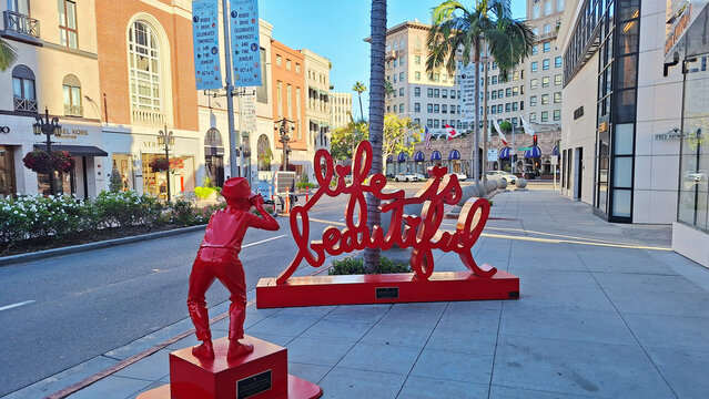 The famous &ldquo;Life is Beautiful&rdquo; text sculpture and photographer statue by artist Mr. Brainwash on the sidewalk of Rodeo Drive, with luxury fashion boutiques. Beverly Hills, California.