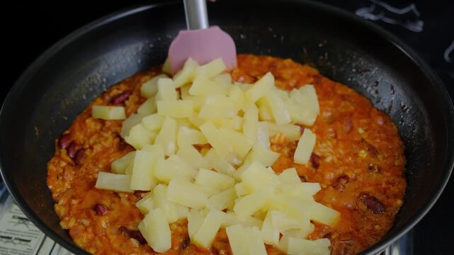 A person stirs a mixture of grains and beans in a black frying pan on a kitchen countertop, adding chopped potato cubes with a pink spatula. Cooking scene.