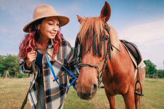 Young woman holding leather reins beside a chestnut horse fitted with decorative bridle fringe outdoors. Equestrian bond concept.