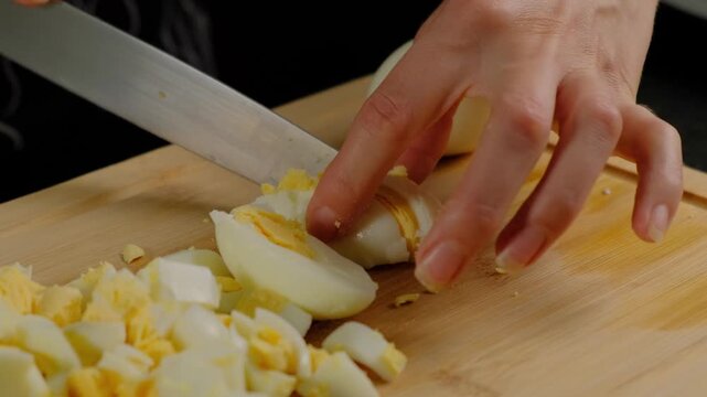Hands in dark sleeves chop hard-boiled eggs with a large knife on a wooden cutting board during cooking in a kitchen, close-up view.