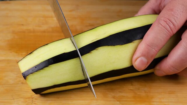 A chef uses a sharp knife to cut a striped peeled eggplant into pieces on a wooden cutting board, preparing healthy organic ingredients with visible flesh and skin textures.