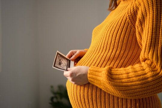 A pregnant woman examines a screening ultrasound photo against a neutral background. The expectant mother viewing a baby scan presents a tender, emotional moment of anticipation awaiting birth.