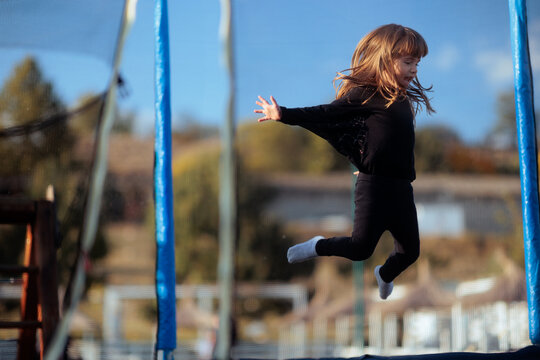 Girl In Bat Costume Jumping High On Outdoor Trampoline. Active child wearing black wings leaps midair pretending to fly during autumn Halloween