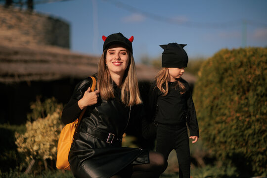 Smiling Mother And Daughter Wearing Black Halloween Costumes Outdoors. Happy parent wearing horned cap dresses her little girl like bat outside