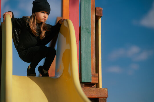 Little Girl Wearing Bat Costume Sitting On Top Of Slide. Happy child dressed in spooky black wings plays at playground during autumn Halloween