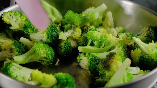 Close-up of hand stir-frying broccoli florets in a metal skillet with spatula at Cooking location, showing browning and caramelization during daytime.