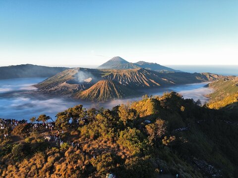 Aerial view of Mount Bromo with its volcanic crater, surrounding sea of sand, and lush hills under a clear blue sky in Tosari, East Java, Indonesia.