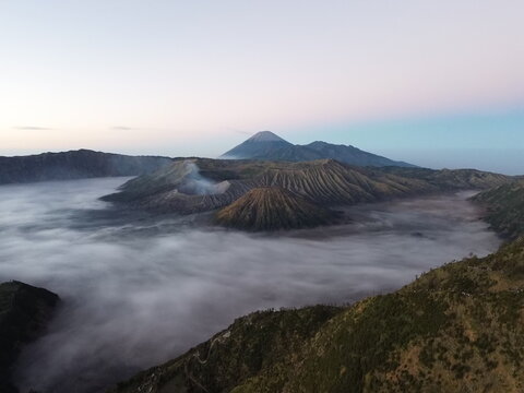 Aerial view of Mount Bromo volcano rising above a sea of clouds with rugged volcanic terrain and soft morning light in Tosari, East Java, Indonesia.