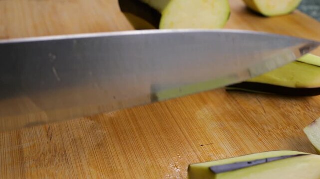 Chef cutting fresh eggplant into thick batons on a wooden board. Close-up view of preparing ingredients for a healthy meal. Culinary process of chopping raw organic vegetables on a table.