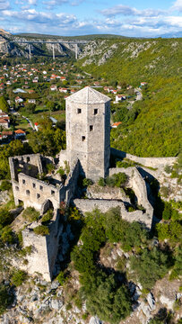 Aerial view of Pocitelj Fortress with its historic stone tower and surrounding ruins, set against a lush green hillside and a distant bridge in Pocitelj, Federation of Bosnia, Bosnia and Herzegovina.