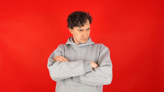 Skeptical young man in grey hoodie with arms crossed looking down away against solid red studio background expressing refusal negative attitude and doubt for advertising.