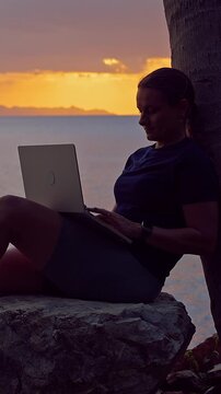 Vertical video. Young Woman Using Laptop While Sitting on a Coastal Rock at Sunset, Remote Work, Digital Nomad Lifestyle, Travel Freedom, Peaceful Productivity