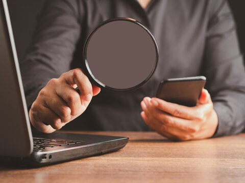 Person holding magnifying glass and smartphone beside laptop