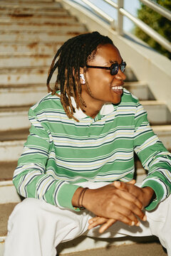 Young Black man with dreadlocks sitting on outdoor stairs, smiling and listening to music with wireless earbuds. Image ideal for youth lifestyle and modern technology marketing, vertical
