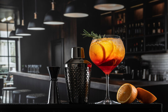 Close-up of an aperol on bar. In tall glass there is bright orange drink with ice and orange slices. Next to it is a stylish black shaker with texture and a jigger
