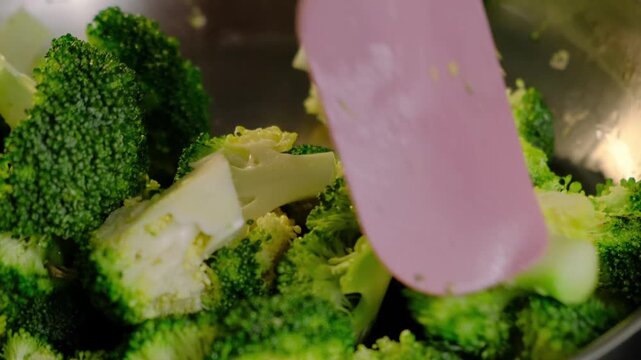 Close-up of cooked broccoli florets and stalks in a metal pot with a pink spatula, set in a kitchen during daytime at Cooking.