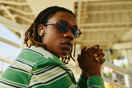 Young adult African American man with dreadlocks wearing sunglasses and wireless earphones outdoors. Image represents youth culture and is suitable for lifestyle marketing, low angle view