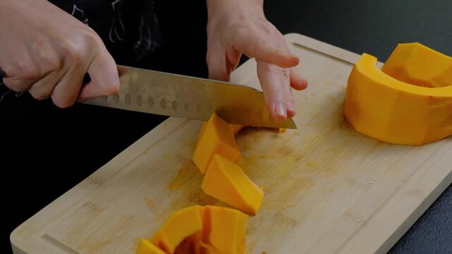 Close-up of hands in dark apron cutting yellow-orange squash with chef's knife on wooden board in Cooking kitchen, daytime.