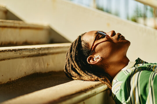 Young adult African American man lying on outdoor stairs listening to music with wireless earphones, representing mental wellbeing. Image suitable for youth lifestyle marketing